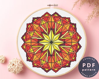 May include: A counted cross stitch pattern featuring a colorful geometric mandala design in shades of red, orange, and yellow. The pattern is displayed in a wooden embroidery hoop.