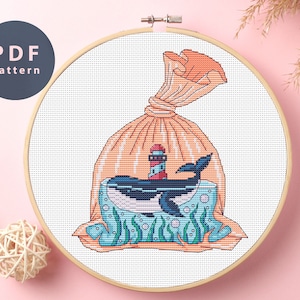 Puede incluir: Un patrón de punto de cruz que presenta una ballena con un faro en su espalda dentro de un saco de arpillera. La ballena es azul y el faro es rojo y blanco. El saco es naranja y tiene un cordón.