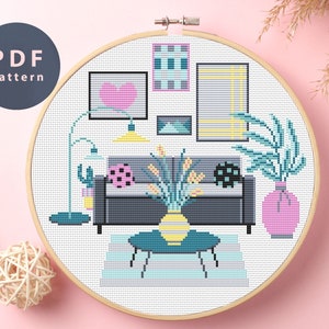 May include: A cross-stitch pattern of a modern living room scene, featuring a gray sofa, artwork, and a coffee table. The design includes a floor lamp, a vase with flowers, and a pink background. The text "PDF pattern" is in a circle.