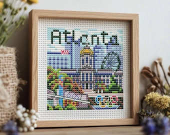 May include: Cross-stitch artwork in a wooden frame featuring the word "Atlanta" above a colorful depiction of the city's skyline, including a gold-domed building and a Ferris wheel. The artwork is displayed on a wooden surface.