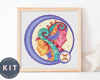 May include: A framed cross-stitch kit featuring the Gemini zodiac sign. The design includes two profiles in shades of pink, yellow, and teal, set against a cream background within a purple circle. The words "COUNTED KIT CROSS-STITCH" are printed on a circular label.
