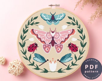 May include: A counted cross stitch pattern featuring a pink and orange moth, two dragonflies, two ladybugs, and white flowers surrounded by green leaves. The pattern is in a round embroidery hoop and the text "COUNTED PDF pattern CROSS STITCH" is on a blue circle.