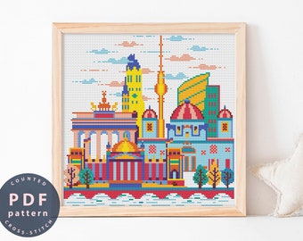 May include: A counted cross stitch pattern featuring a colorful cityscape with buildings, a tower, and trees. The pattern is framed in a light wood frame.