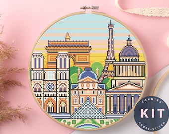 May include: A counted cross stitch kit featuring a colorful pixelated design of famous Parisian landmarks, including the Eiffel Tower, Notre Dame Cathedral, and the Arc de Triomphe. The kit includes a printed pattern and embroidery floss. The text "COUNTED KIT CROSS STITCH" is printed on a blue circle.