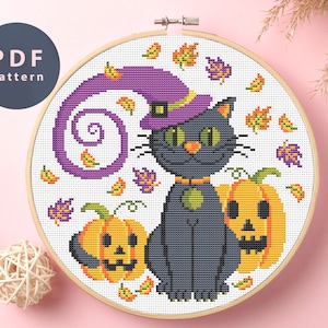 May include: Cross stitch pattern featuring a black cat wearing a purple witch hat and holding a jack-o'-lantern. The design is surrounded by orange and purple leaves.