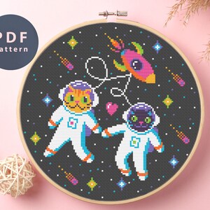 Könnte beinhalten: Kreuzstichmuster mit zwei Katzen, die als Astronauten im Weltraum verkleidet sind. Die Katzen tragen weiße Raumanzüge mit blauen Akzenten und Helmen. Sie schweben im Weltraum mit einem Raumschiff und Sternen. Das Muster ist in einem Schwarz-Weiß-Farbschema mit bunten Akzenten gehalten.