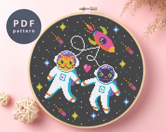 May include: Cross stitch pattern featuring two cats dressed as astronauts in space. The cats are wearing white spacesuits with blue accents and helmets. They are floating in space with a rocket ship and stars. The pattern is in a black and white color scheme with colorful accents.
