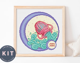 May include: A framed cross-stitch kit featuring the Aquarius zodiac symbol. The design includes a pink and red water vessel pouring turquoise water, set against a light yellow background within a purple circle. The words "COUNTED KIT CROSS-STITCH" are on a circular label.