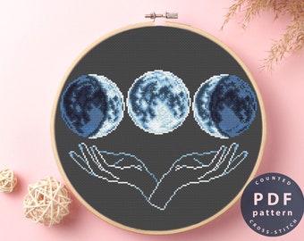 May include: Counted cross stitch pattern PDF for a moon phases design. The pattern features three moons in different phases, with a pair of hands holding them up. The background is dark gray.