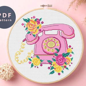 May include: Cross stitch pattern featuring a pink rotary phone with a yellow dial and floral accents. The phone has a cord with a curly end. The pattern is surrounded by a white background.