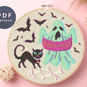 Puede incluir: Patrón de punto de cruz con un fantasma verde con el texto "Boo to you!" en una pancarta rosa, un gato negro y murciélagos sobre un fondo beige.