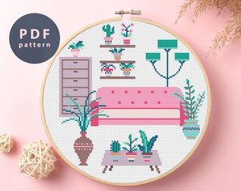 May include: Cross stitch pattern of a living room with a pink sofa, a dresser, a lamp, and plants in pots. The pattern is in a white embroidery hoop.