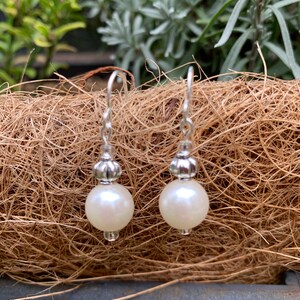 Può includere: Un paio di orecchini pendenti con perle. Ogni orecchino presenta una grande perla rotonda bianco sporco sospesa a un gancio in metallo color argento. Il gancio è collegato a una perlina decorativa argentata. Lo sfondo è un materiale marrone intrecciato.