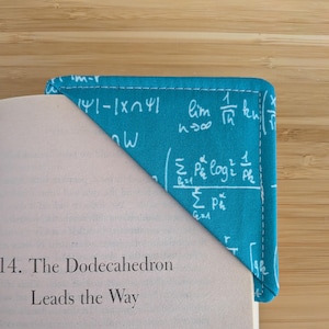 May include: A teal fabric corner bookmark with white mathematical equations and symbols is placed on the corner of a book. The bookmark is stitched with white thread. The book's page has the title "The Dodecahedron Leads the Way."