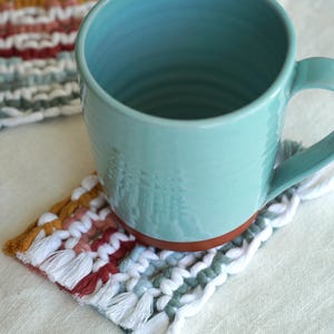 Könnte beinhalten: Eine türkisfarbene Keramiktasse steht auf einem handgefertigten Makramee-Untersetzer. Der Untersetzer hat ein gewebtes Design mit weißen, roten, gelben und grünen Fäden und Fransen. Ein weiterer Untersetzer ist im Hintergrund zu sehen.