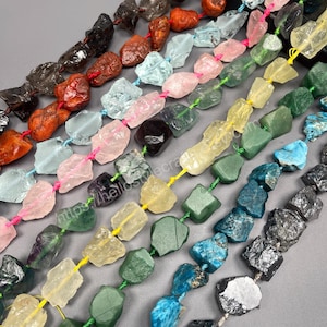Puede incluir: Hileras de cuentas de piedras preciosas en bruto de varios colores, incluyendo rosa, verde, amarillo y azul. Las cuentas están ensartadas con cordones coloridos, creando una exhibición visualmente atractiva de componentes de joyería de piedra natural.