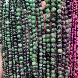 May include: A close-up of multiple strands of green, black, and pink gemstone beads. The beads are round and have a speckled appearance.