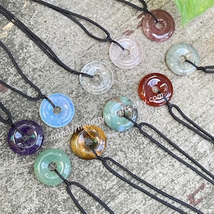 May include: A selection of gemstone donut pendants in diverse colours, such as purple, blue, green, and brown, each on a black cord. The pendants are circular with a central hole. The image includes the text "https://hellostylecrafts.etsy.com".