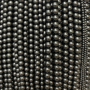 May include: A close-up of a large quantity of black pearl beads. The beads are arranged in rows and are all the same size and shape.