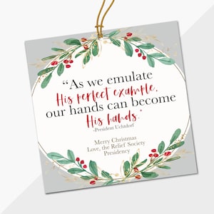 May include: A gray Christmas ornament with a gold wreath and red berries. The ornament features a quote from President Uchtdorf: "As we emulate His perfect example, our hands can become His hands."