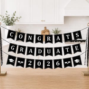May include: A black banner with white text that reads "CONGRATS GRADUATE 2026". The banner is strung between two gold-colored stands, and is decorated with graduation cap and diploma icons. Black and white beads hang from the stands.