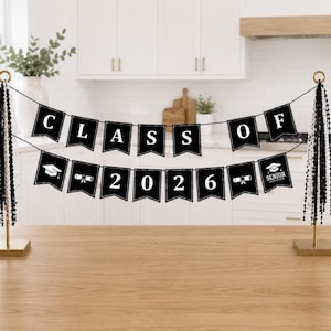 May include: A black banner with white text that reads "CLASS OF 2026" is displayed between two gold stands. The banner has a graduation cap and diploma graphic. Black and white beads hang from the stands.