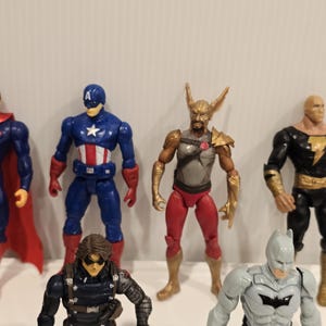 May include: A collection of action figures featuring various superheroes. The figures are brightly colored and detailed, including Captain America in blue and red, and Batman in gray. The figures are standing on a white surface.