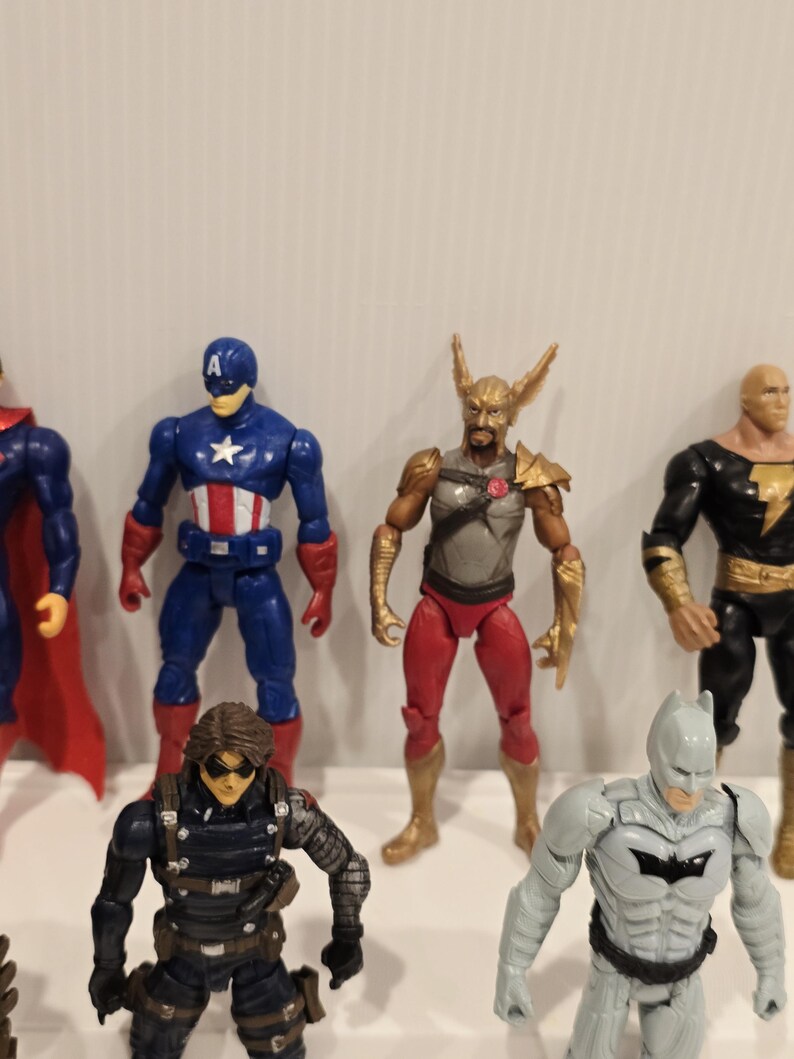 May include: A collection of action figures featuring various superheroes. The figures are brightly colored and detailed, including Captain America in blue and red, and Batman in gray. The figures are standing on a white surface.