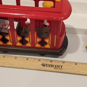 May include: A red plastic toy trolley with yellow and brown accents. The trolley has a rectangular shape with open sides and a curved roof. The trolley is sitting on a brown base with black wheels. A wooden ruler is next to the trolley.