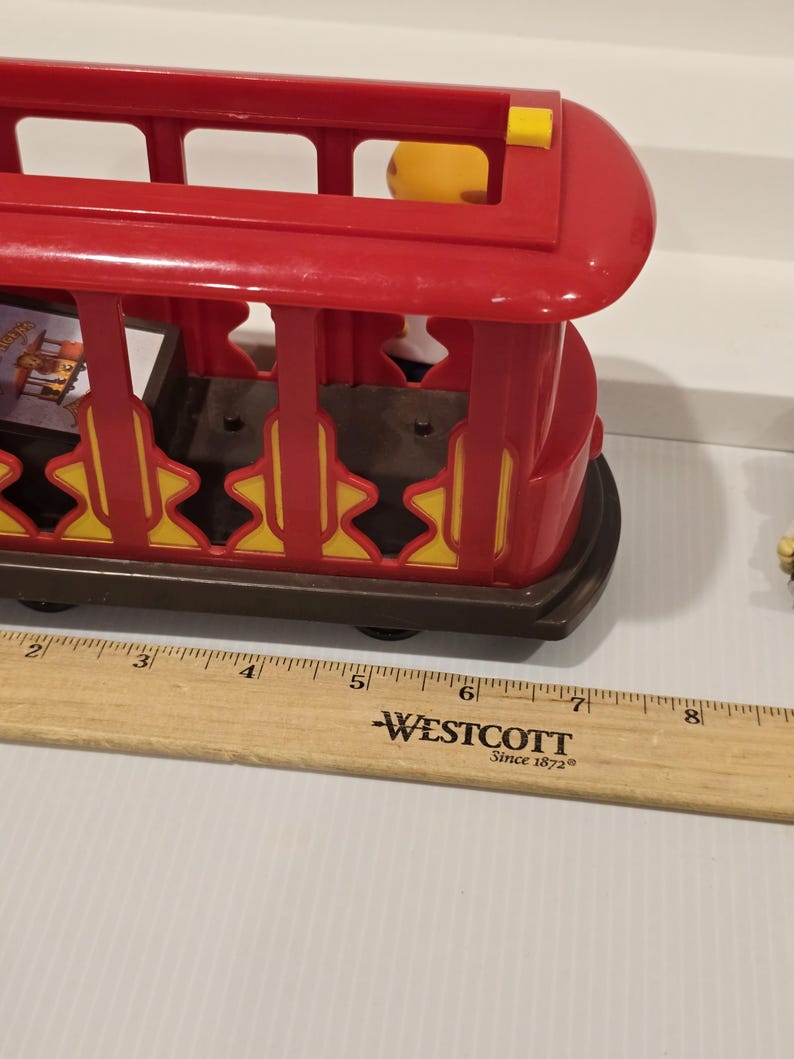 May include: A red plastic toy trolley with yellow and brown accents. The trolley has a rectangular shape with open sides and a curved roof. The trolley is sitting on a brown base with black wheels. A wooden ruler is next to the trolley.