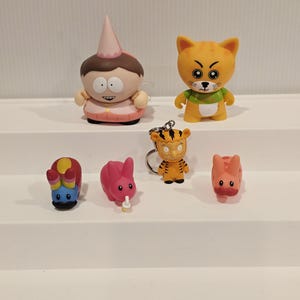 May include: Collection of colorful figurines, including a character with a pink hat and outfit, an orange cat, a tiger keychain, and several small animal figures. The figurines are arranged on a white surface.