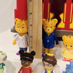 K&ouml;nnte beinhalten: Eine Sammlung farbenfroher Daniel Tiger's Neighborhood-Figuren, darunter Daniel Tiger, Katerina Kittycat und O die Eule. Die Figuren sind etwa 7,6 cm gro&szlig;. Im Hintergrund befindet sich ein roter und gelber Spielzeugzug.