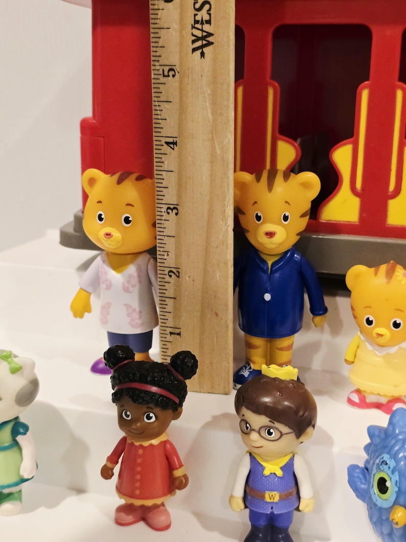 K&ouml;nnte beinhalten: Eine Sammlung farbenfroher Daniel Tiger's Neighborhood-Figuren, darunter Daniel Tiger, Katerina Kittycat und O die Eule. Die Figuren sind etwa 7,6 cm gro&szlig;. Im Hintergrund befindet sich ein roter und gelber Spielzeugzug.