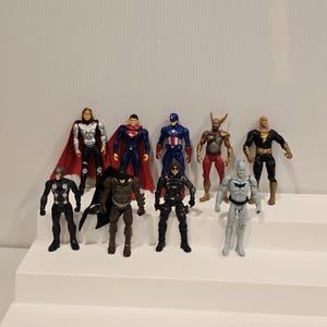 May include: A collection of superhero action figures, including Thor, Superman, Captain America, and Batman, are displayed on a white stepped platform. The figures are in various poses and costumes, with vibrant colors and detailed designs.