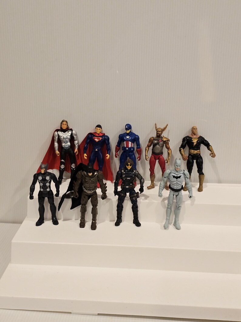 May include: A collection of superhero action figures, including Thor, Superman, Captain America, and Batman, are displayed on a white stepped platform. The figures are in various poses and costumes, with vibrant colors and detailed designs.