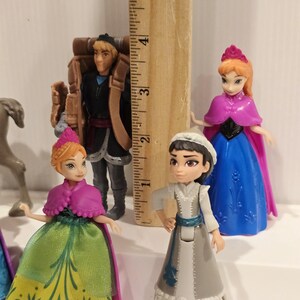 May include: A collection of miniature figurines featuring characters from the animated film "Frozen." The figures include Anna, Elsa, Kristoff, and other characters. The figurines are colorful and detailed, with a ruler in the background for scale.