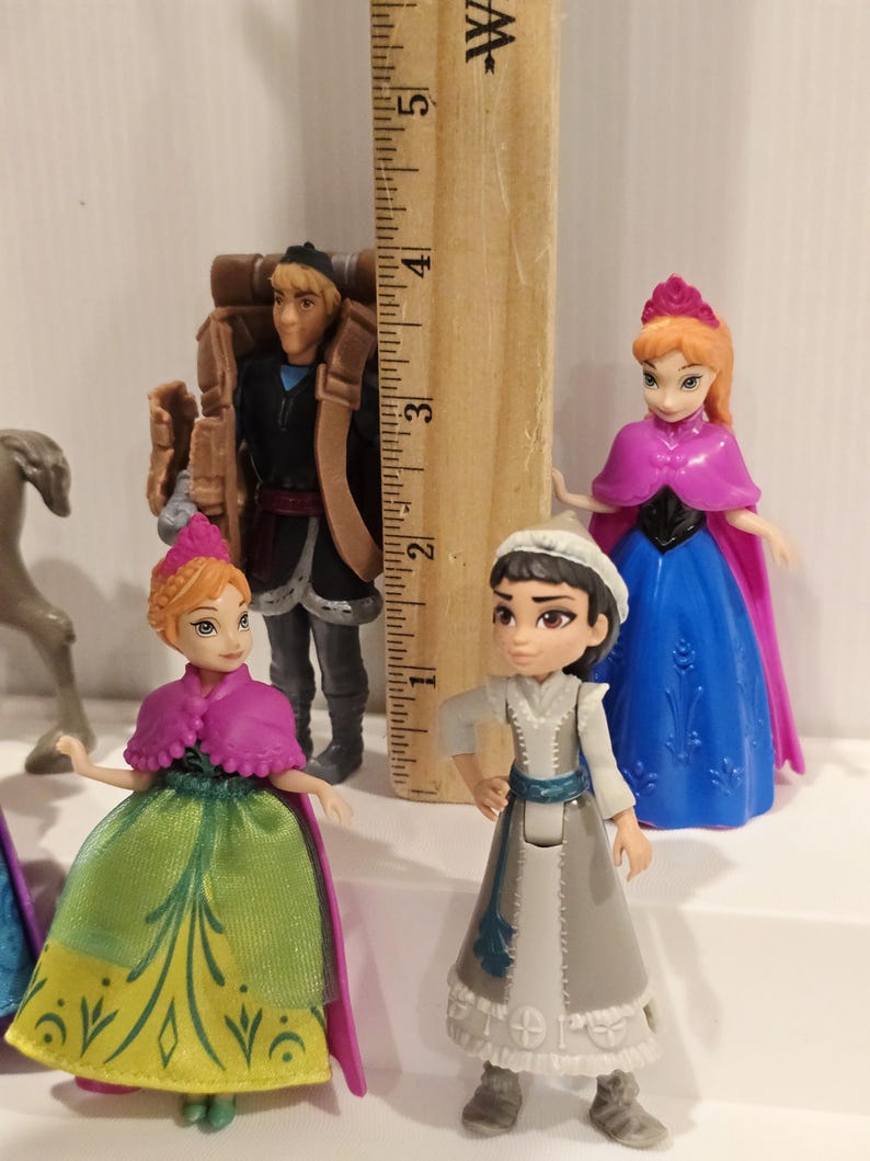 May include: A collection of miniature figurines featuring characters from the animated film "Frozen." The figures include Anna, Elsa, Kristoff, and other characters. The figurines are colorful and detailed, with a ruler in the background for scale.