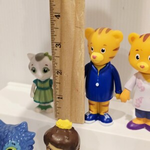 May include: A collection of plastic character figurines including a small grey cat in a green dress and a tiger in a blue jacket. A wooden ruler is positioned vertically next to the tiger, showing a height of approximately 3.5 inches.