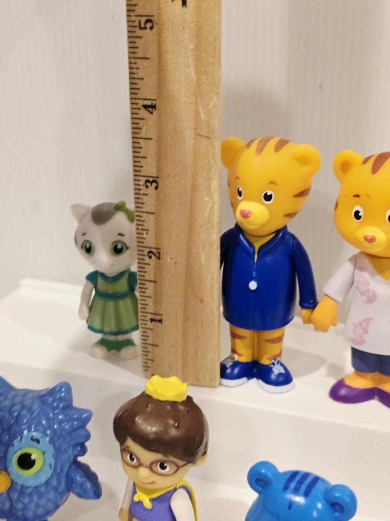 May include: A collection of plastic character figurines including a small grey cat in a green dress and a tiger in a blue jacket. A wooden ruler is positioned vertically next to the tiger, showing a height of approximately 3.5 inches.