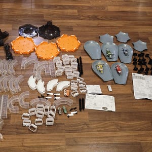 May include: A collection of toy race car track components, including clear plastic track pieces, orange and white track supports, and gray car holders. Also included are small toy cars in various colors, black connectors, and instruction manuals. The items are arranged on a wooden surface.