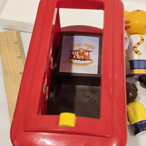 May include: A red plastic toy bus with yellow accents. The bus has an open top and a rectangular window displaying a graphic with the text "DANIEL TIGER'S NEIGHBORHOOD". Other toy figures are visible in the background.