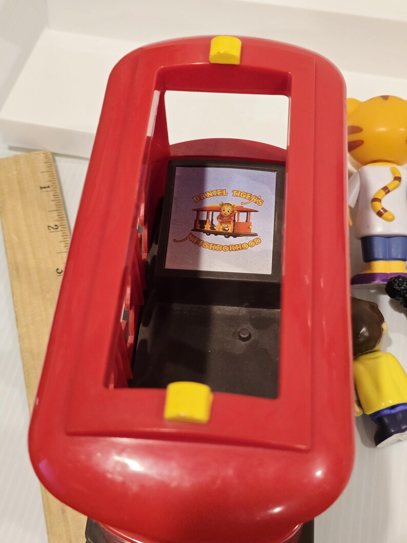May include: A red plastic toy bus with yellow accents. The bus has an open top and a rectangular window displaying a graphic with the text "DANIEL TIGER'S NEIGHBORHOOD". Other toy figures are visible in the background.
