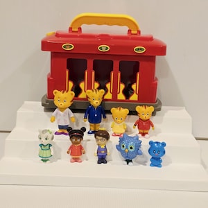 K&ouml;nnte beinhalten: Ein roter Spielzeug-Zugwagen mit gelbem Griff, zusammen mit mehreren kleinen Figuren. Die Figuren zeigen Charaktere in verschiedenen Outfits, darunter ein Arztkittel, ein blauer Anzug und eine rote Jacke. Das Spielzeug ist auf einer wei&szlig;en, abgestuften Oberfl&auml;che angeordnet.