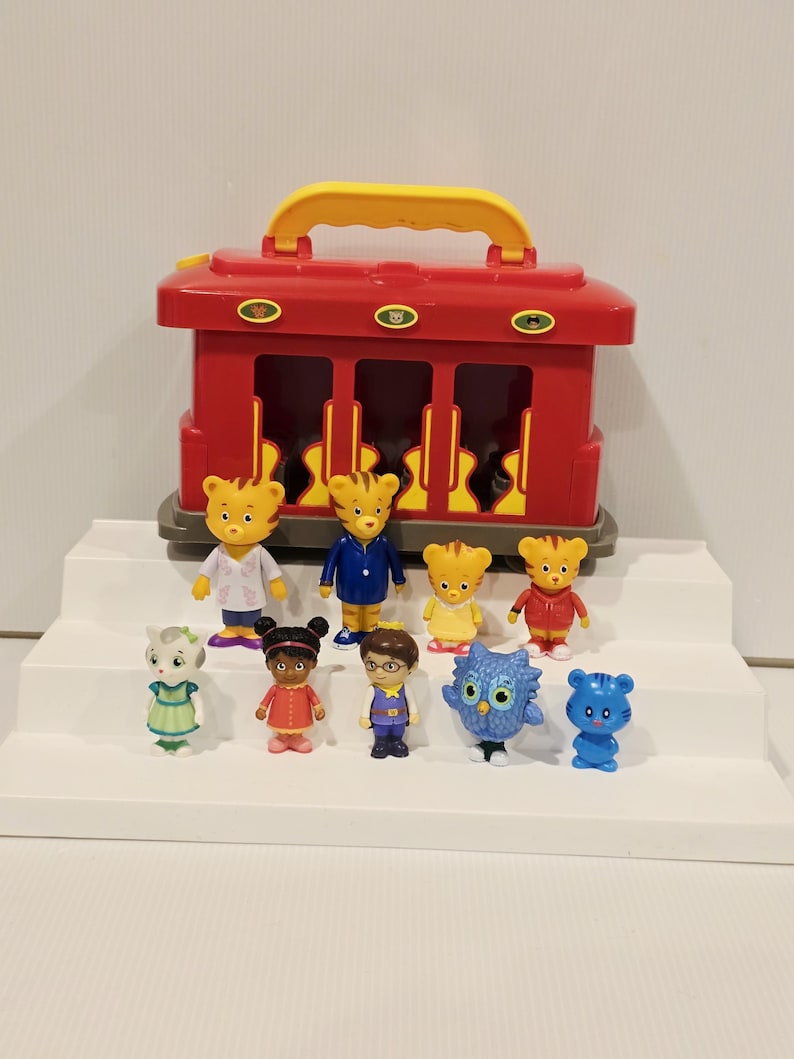 K&ouml;nnte beinhalten: Ein roter Spielzeug-Zugwagen mit gelbem Griff, zusammen mit mehreren kleinen Figuren. Die Figuren zeigen Charaktere in verschiedenen Outfits, darunter ein Arztkittel, ein blauer Anzug und eine rote Jacke. Das Spielzeug ist auf einer wei&szlig;en, abgestuften Oberfl&auml;che angeordnet.
