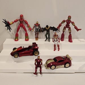 May include: A collection of superhero action figures and toy cars. The figures are in various poses and feature red, gold, and black color schemes. The toy cars are primarily red with gold accents.