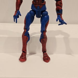 Pode incluir: Uma figura de a&ccedil;&atilde;o do Homem-Aranha, predominantemente vermelha e azul, com padr&otilde;es de teia de aranha detalhados. A figura est&aacute; em pose de p&eacute;, mostrando seus membros articulados e o design ic&ocirc;nico do traje. A figura tem aproximadamente 15 cm de altura.