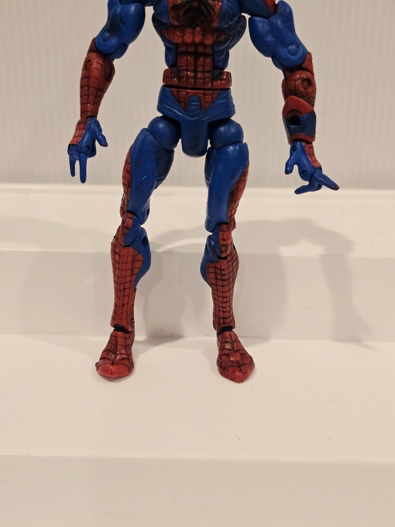 Pode incluir: Uma figura de a&ccedil;&atilde;o do Homem-Aranha, predominantemente vermelha e azul, com padr&otilde;es de teia de aranha detalhados. A figura est&aacute; em pose de p&eacute;, mostrando seus membros articulados e o design ic&ocirc;nico do traje. A figura tem aproximadamente 15 cm de altura.