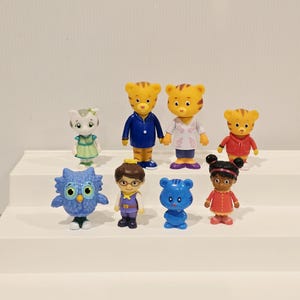 May include: A collection of eight small plastic character figurines. The set includes two adult tigers, a child tiger, a white cat, a blue owl, a human child with glasses, a small blue tiger, and a human child with dark hair in two buns.