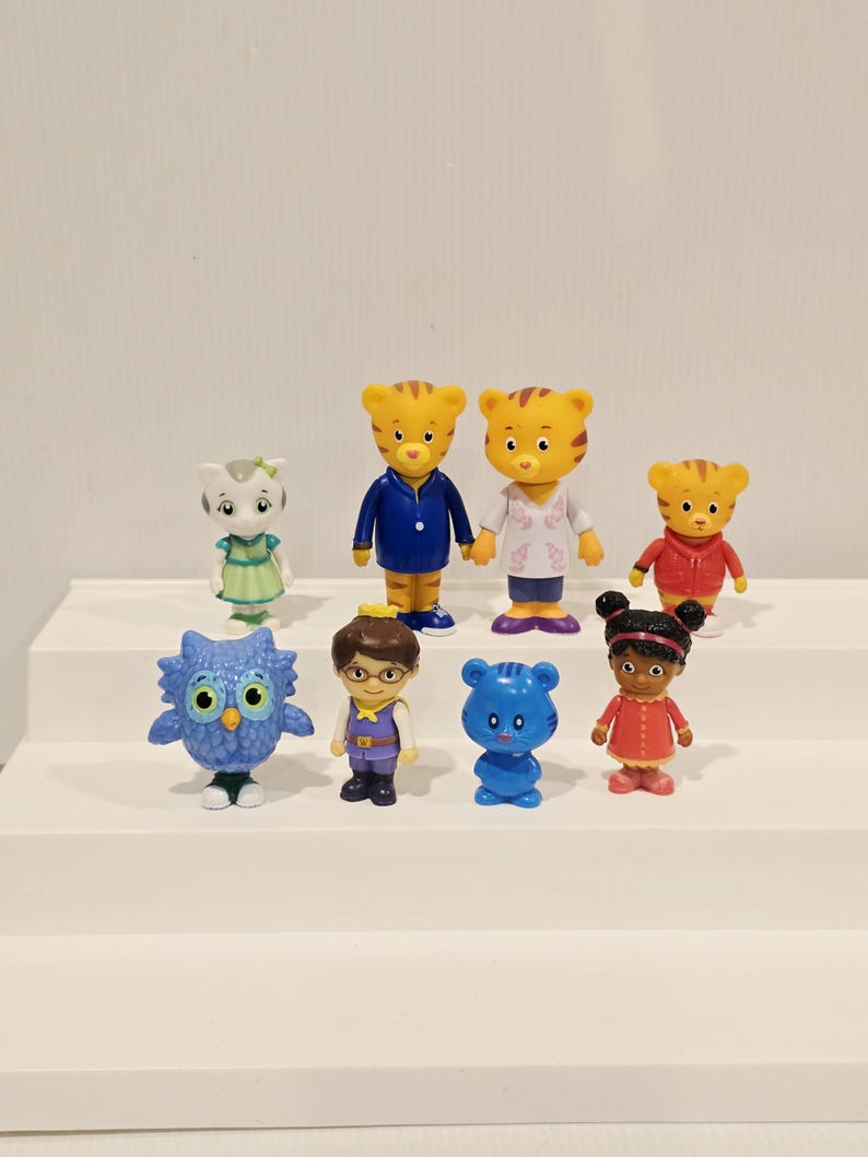 May include: A collection of eight small plastic character figurines. The set includes two adult tigers, a child tiger, a white cat, a blue owl, a human child with glasses, a small blue tiger, and a human child with dark hair in two buns.