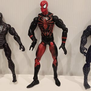 May include: A red and black Spider-Man action figure with a spider emblem on the chest. The figure has articulated joints and is posed with arms extended. Other Spider-Man figures are visible in the background.